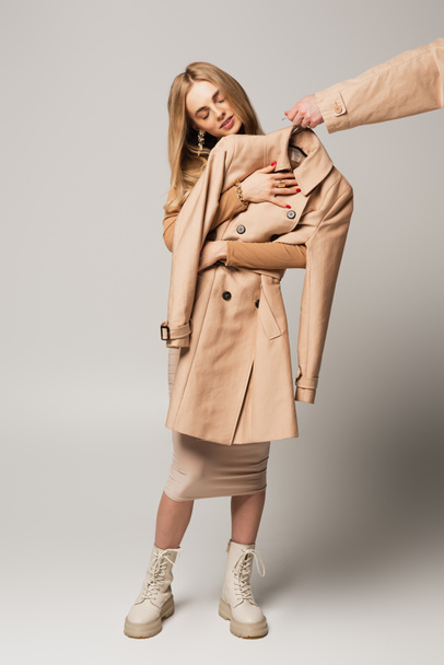 jonge vrouw houden jas in de buurt hand met hanger op grijze achtergrond - Foto, afbeelding