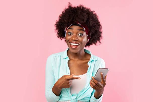 Волнующая африканская женщина указывает пальцем на смартфон на розовом фоне - Фото, изображение