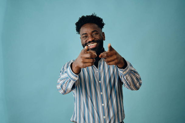 portrait of a young african american man with a beard smiling and pointing at camera and a blue shirt on a gray background - Photo, Image