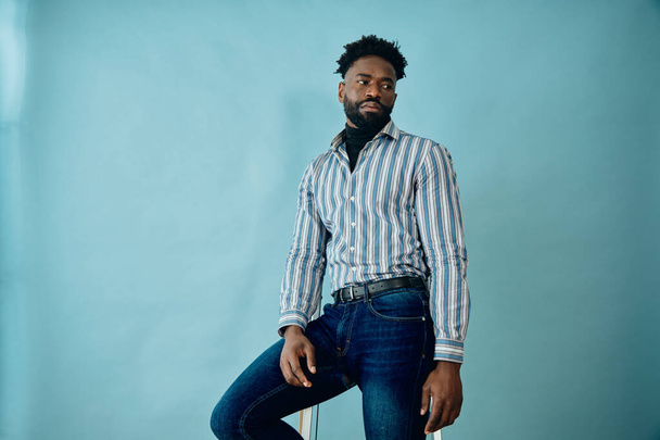 portrait of a handsome african american man in a blue and white stripe shirt and jeans on a blue background - Photo, Image