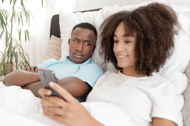 У молодой пары проблемы. Ревнивый муж смотрит, как его жена использует мобильный телефон на кровати дома - Фото, изображение