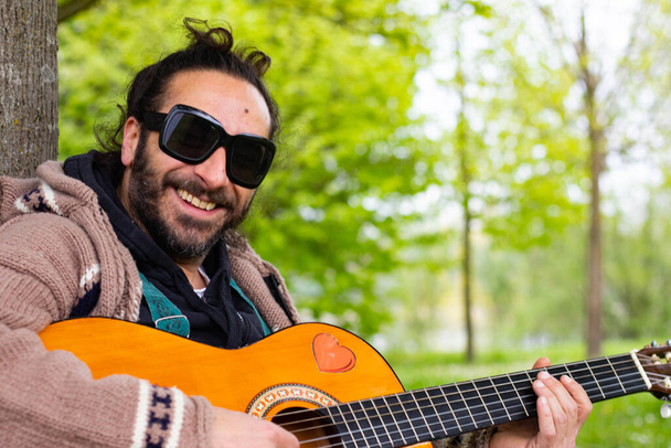 Hippy man with beard and glasses playing guitar outdoors on the grass - Photo, Image