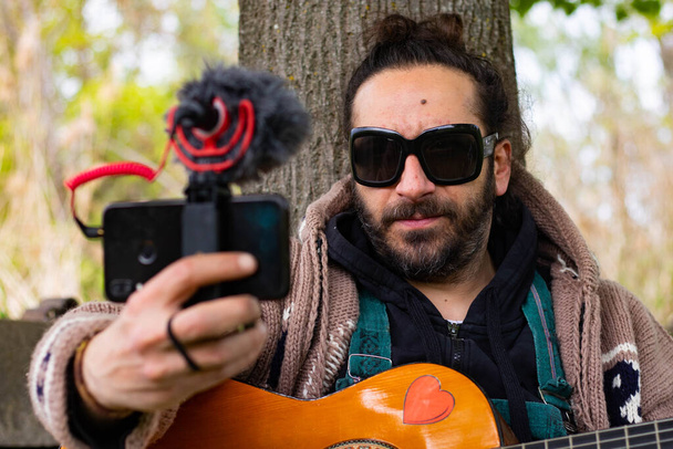 Hippy άνθρωπος με γενειάδα και γυαλιά που παίζουν κιθάρα κάνοντας ένα βίντεο τον εαυτό της σε εξωτερικούς χώρους στο γρασίδι - Φωτογραφία, εικόνα