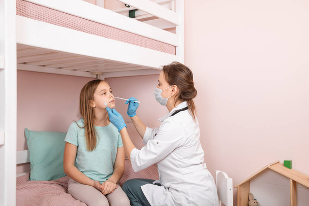 Pediatrician taking nasal mucus test sample from elementary age girl's nose performing respiratory virus testing procedure at home during coronavirus COVID-19 pandemic quarantine - 写真・画像