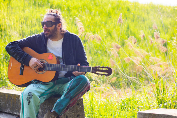 Хиппи с бородой и в очках играет на гитаре на траве - Фото, изображение