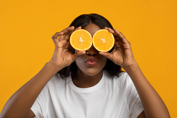 Funny african american lady covering eyes with orange halves - Fotó, kép