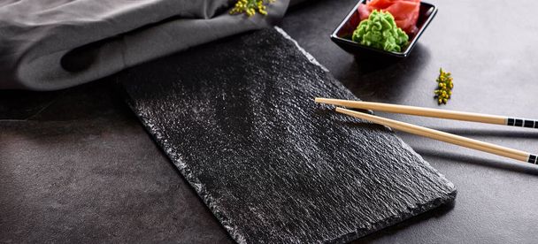 Ασιατική κουζίνα - Black Slate Plate με Chopstick. Τοποθετήστε τη ρύθμιση με wasabi και πίκλα τζίντζερ. Κενό πιάτο σούσι σε μαύρο πέτρινο τραπέζι - Φωτογραφία, εικόνα