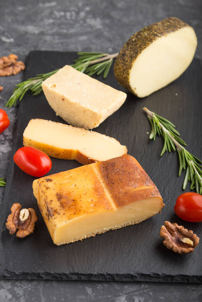 Smoked cheese and various types of cheese with rosemary and tomatoes on black slate board on a black concrete background. Side view, close up, selective focus. - Photo, Image