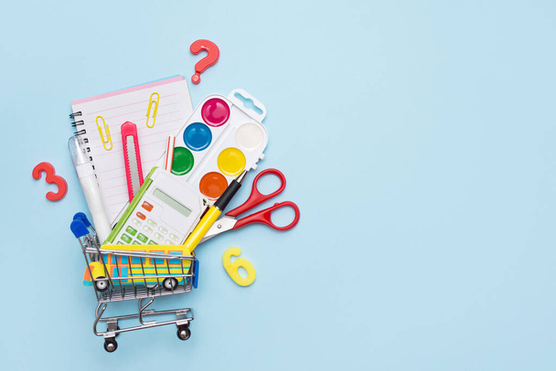 Schulbedarf: Bleistifte, Pastelle, Lineale und Notizbuch im Einkaufswagen, auf blauem Hintergrund mit Kopierraum - Foto, Bild