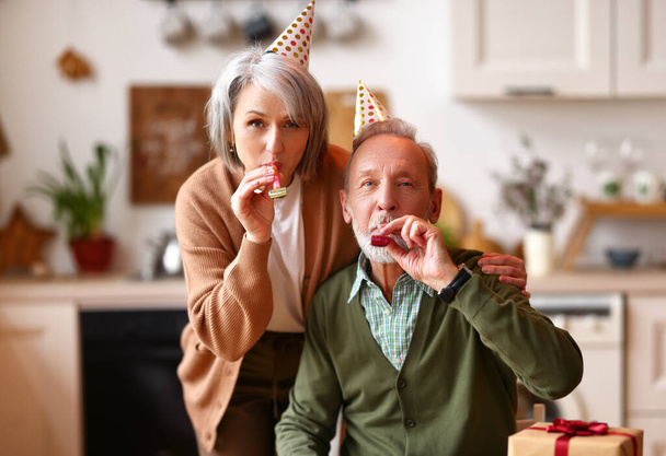 Onnellinen kaunis vanhempi pari juhlii syntymäpäivää kotona, iäkäs mies ja nainen yllään puolue hatut syleilee, puhaltaa pillit ja katsot kameraa. Juhla ja loma käsite - Valokuva, kuva