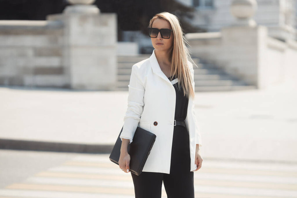 Street style portrait of serious business woman in a sunglasses, wearing white jacket and black trousers, walking along the city, working with laptop  - Фото, зображення
