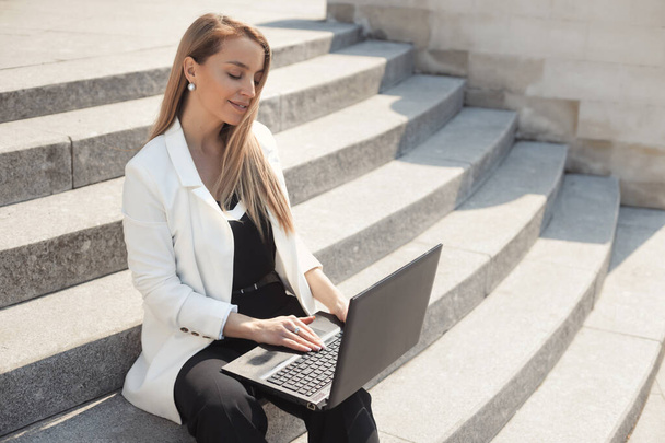 Street style portrait of serious business woman, wearing white jacket and black trousers, walking along the city, working with laptop sitting on steps - Foto, afbeelding
