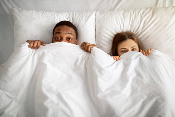 Couple interracial ludique se cachant sous une couverture blanche au lit, vue sur le dessus - Photo, image