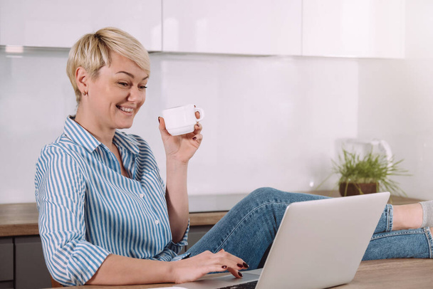 Wesoła młoda kobieta pije kawę i rozmawia na wideokonferencji na laptopie siedząc w domu - Zdjęcie, obraz