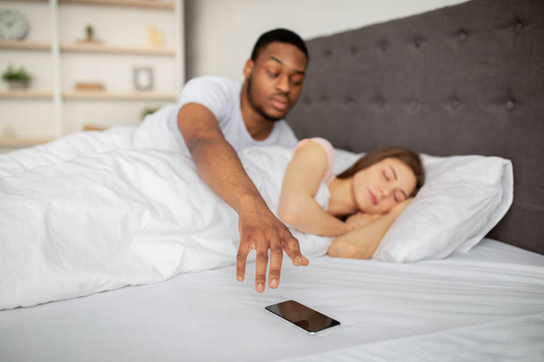 Verdachte zwarte man die thuis zijn slapende vrouw zijn mobiele telefoon checkt. Relatieproblemen - Foto, afbeelding