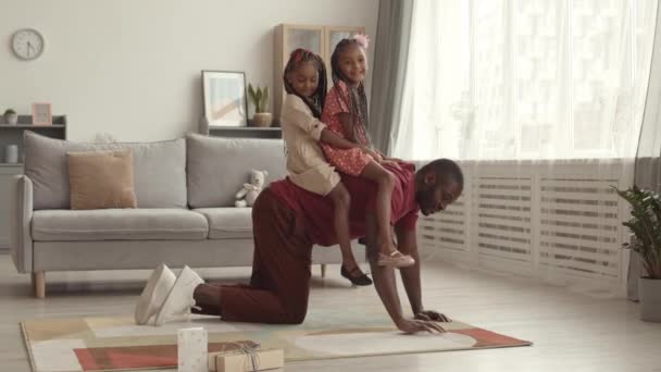 Breed shot van jonge Afrikaanse vader lopen op handen en voeten in de woonkamer, paardrijden twee kleine schattige vrouwelijke kinderen op zijn rug, glimlachen - Video