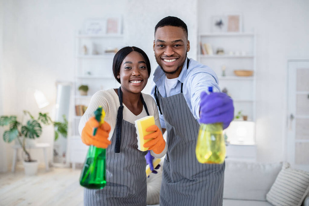 Szczęśliwy czarny facet i jego żona celują detergenty spray przed kamerą, wykonując domowe sprzątanie, w pomieszczeniach - Zdjęcie, obraz