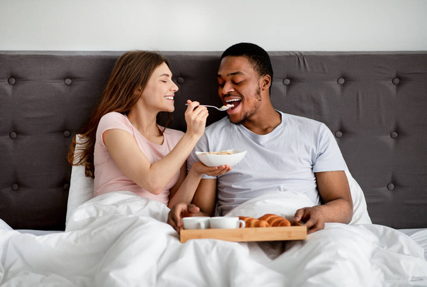Heureux couple multiracial prenant le petit déjeuner au lit, jeune femme nourrissant son mari avec de délicieuses céréales - Photo, image