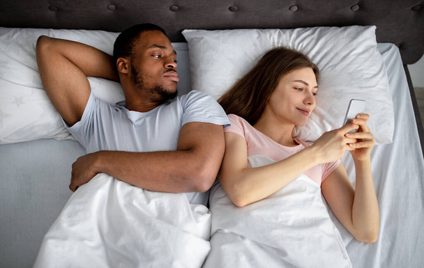 Irritated black guy feeling jealous of his girlfriend stuck in smartphone in bed, top view. Phubbing concept - Фото, изображение