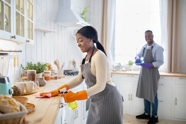 Веселая черная женщина убирает кухонный стол, ее парень моет посуду на заднем плане, пустое место - Фото, изображение