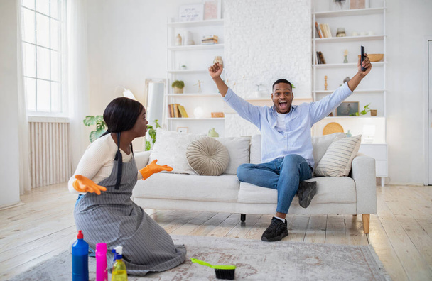 Podekscytowany czarny facet oglądający futbol w telewizji, szczęśliwy po zwycięstwie swojej drużyny, wściekły obrażony sprzątający dom sam - Zdjęcie, obraz
