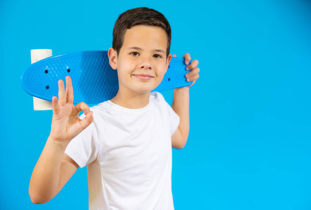Молодий хлопчик тримає скейтборд зі знаком добре ізольовано на синьому фоні
. - Фото, зображення