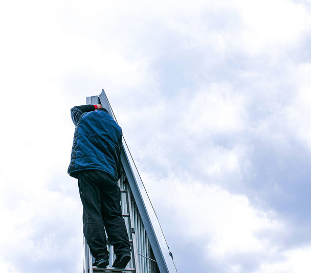 曇りの日に屋外広告バナーメカニズムの修復.専門家は階段を登って看板に向かいます。リスクの高い仕事だ。仕事の高さ. - 写真・画像