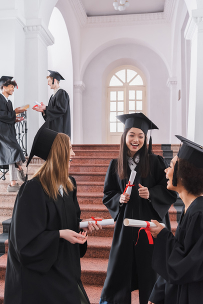 Estudiantes multiétnicos sonrientes con diplomas hablando en la universidad  - Foto, Imagen