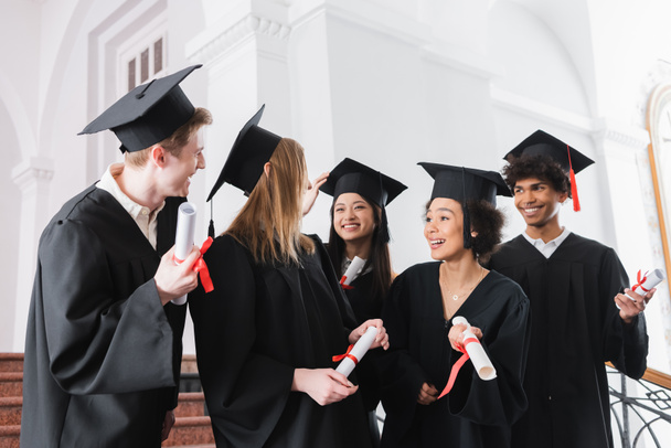 Positive multiethnic graduates looking at friends with diplomas  - Fotografie, Obrázek