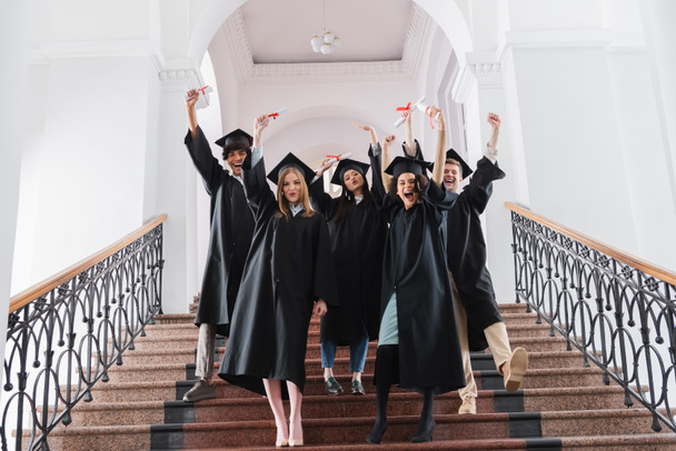 Excited multiethnic graduates with diplomas standing on stairs in university hall - 写真・画像