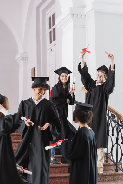 Estudante alegre apontando para diploma perto de amigos multiculturais em primeiro plano turvo  - Foto, Imagem