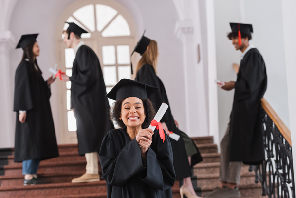 Happy african american graduate holding diploma near blurred friends in university  - Photo, Image