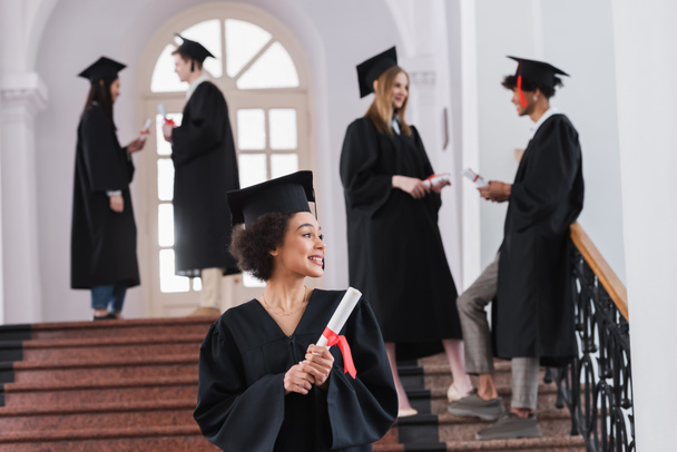 Sorrindo afro-americano graduado com diploma de pé perto de amigos em fundo turvo  - Foto, Imagem