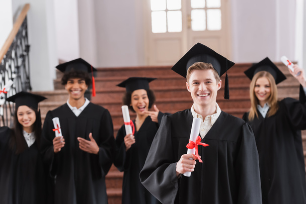 Улыбающийся студент в кепке держит диплом рядом с межрасовыми друзьями на размытом фоне  - Фото, изображение