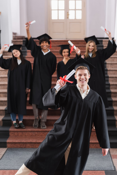 Glücklicher Absolvent mit Diplom in der Nähe von interrassischen Freunden in akademischen Kleidern  - Foto, Bild
