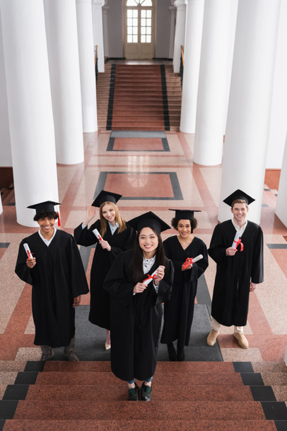 Blick aus der Vogelperspektive auf lächelnde multiethnische Junggesellen mit Diplomen auf der Treppe der Universität  - Foto, Bild