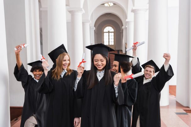 Aziatisch afgestudeerd met diploma tonen ja gebaar in de buurt opgewonden vrienden  - Foto, afbeelding