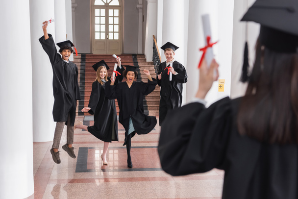 Cheerful multiethnic graduates holding diplomas near friend on blurred foreground  - Fotografie, Obrázek