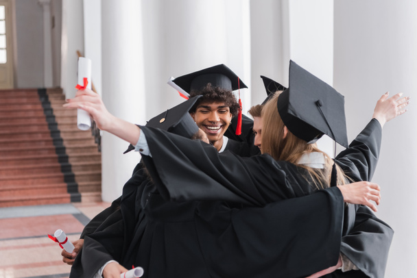Sonriente afroamericano graduado con diploma abrazando amigos  - Foto, imagen