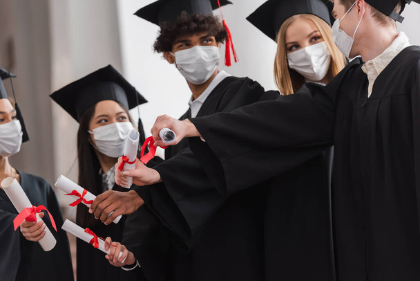 Multicultural bachelors in medical masks on blurred background holding diplomas  - Photo, Image