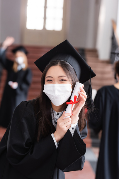 Akademik elbise ve tıp maskesi içinde diplomasını kurdeleyle tutan Asyalı bir mezun.  - Fotoğraf, Görsel