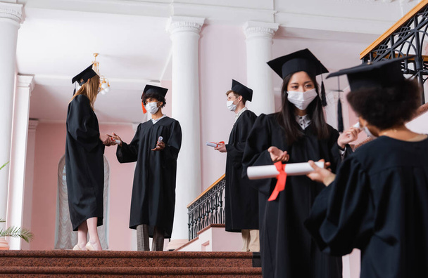 Interrassische Studenten in medizinischen Masken und akademischen Kleidern sprechen in der Aula der Universität  - Foto, Bild