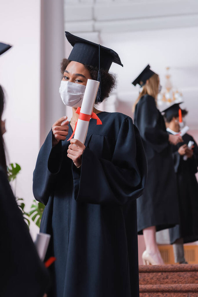Estudiante afroamericano en máscara médica señalando diploma  - Foto, imagen