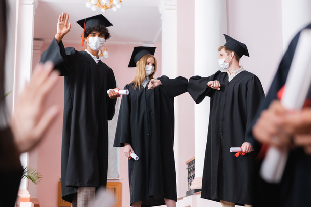 African american graduate in medical mask waving hand near friends doing embow bump  - Photo, Image