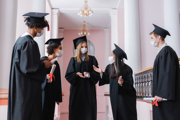 Asian graduate in protective mask talking to interracial friends with diplomas  - Photo, Image