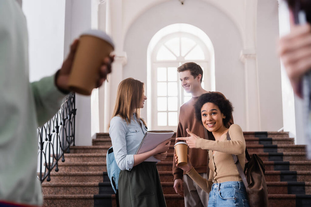 Smiling african american student with coffee to go pointing near friends in university  - Photo, Image