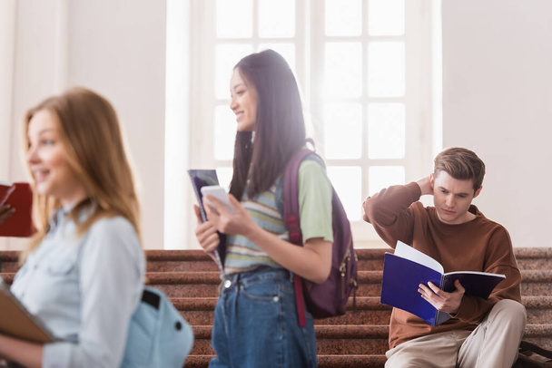 Estudiante mirando cuaderno cerca de personas multiétnicas en primer plano borrosa  - Foto, Imagen