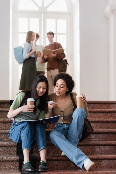 Interraciale studenten met koffie te gaan, notebook en smartphone zitten op de trap  - Foto, afbeelding