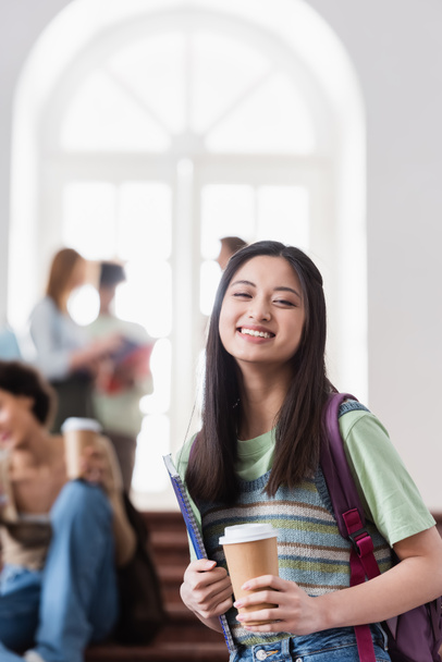 Sonriendo asiática estudiante con papel taza y portátil mirando a la cámara  - Foto, imagen