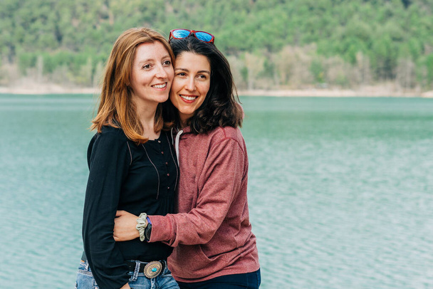 Brunette woman and red-haired woman hiking in a lake with mountains in the background - Фото, изображение